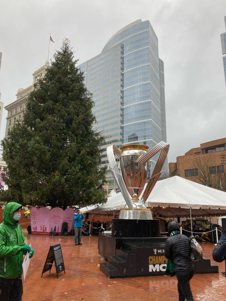A giant replica of the MLS Cup, on display at Portland's Pioneer Courthouse Square, with the Square's Christmas Tree in the background.